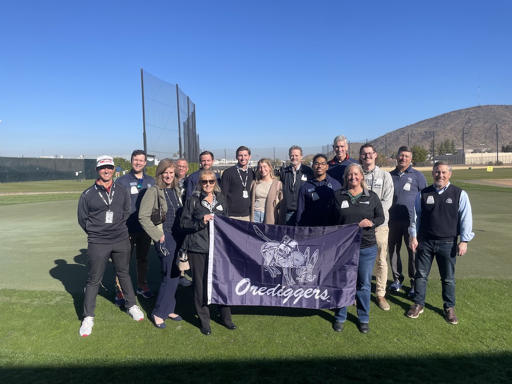 A group of Mines alumni stand on a golf course at the PING headquarters holding an Oredigger flag