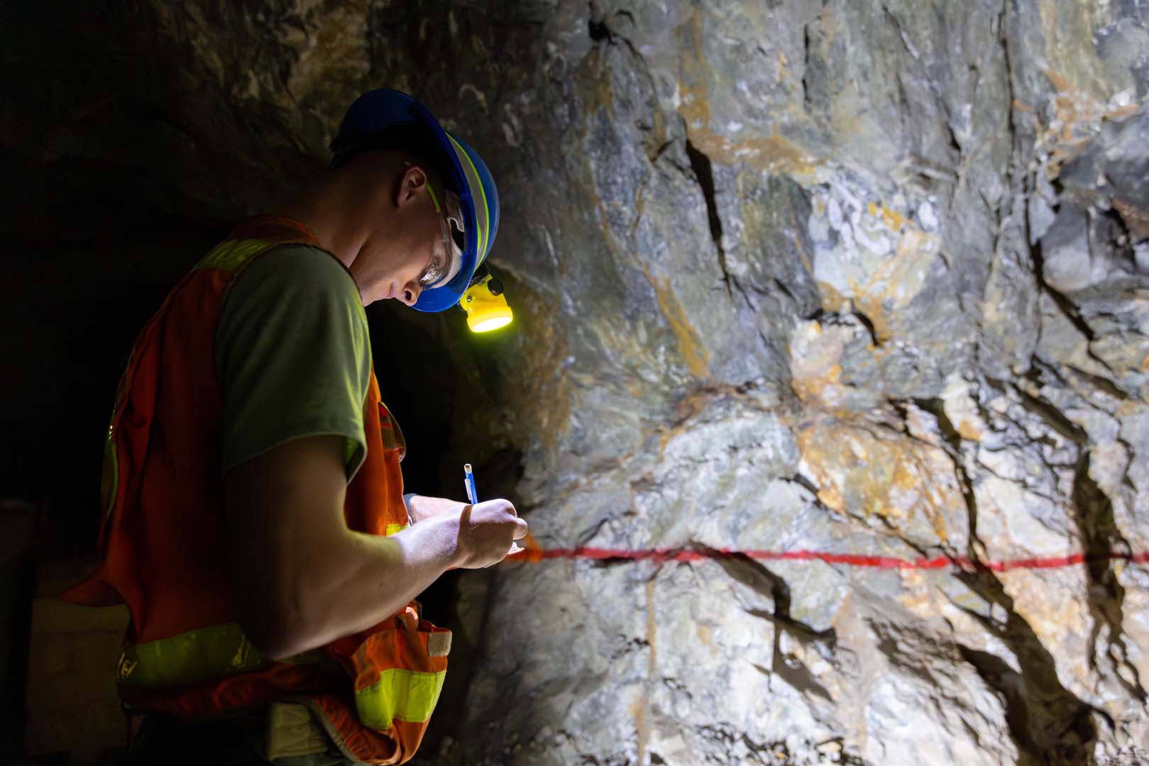 Mines student wears a safety vest and hard hat within Edgar Mine. He is taking notes while standing in front of a rock face.